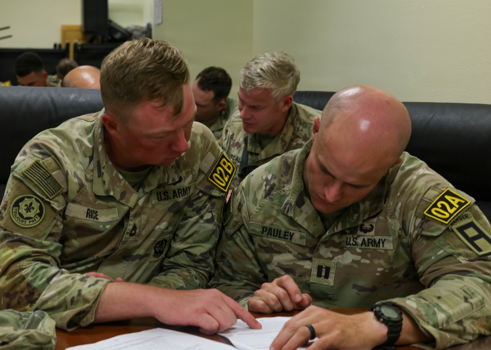 Two U.S. Army soldiers in OCP (Operational Camouflage Pattern) uniforms are seated at a table reviewing documents together. The soldier on the left is identified by name tape as 'RICE' wearing team marker '02B', while the soldier on the right is identified as 'PAULEY' wearing team marker '02A' and captain rank insignia. Additional soldiers are visible in the background in a similar indoor military setting.