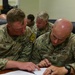 Two U.S. Army soldiers in OCP (Operational Camouflage Pattern) uniforms are seated at a table reviewing documents together. The soldier on the left is identified by name tape as 'RICE' wearing team marker '02B', while the soldier on the right is identified as 'PAULEY' wearing team marker '02A' and captain rank insignia. Additional soldiers are visible in the background in a similar indoor military setting.