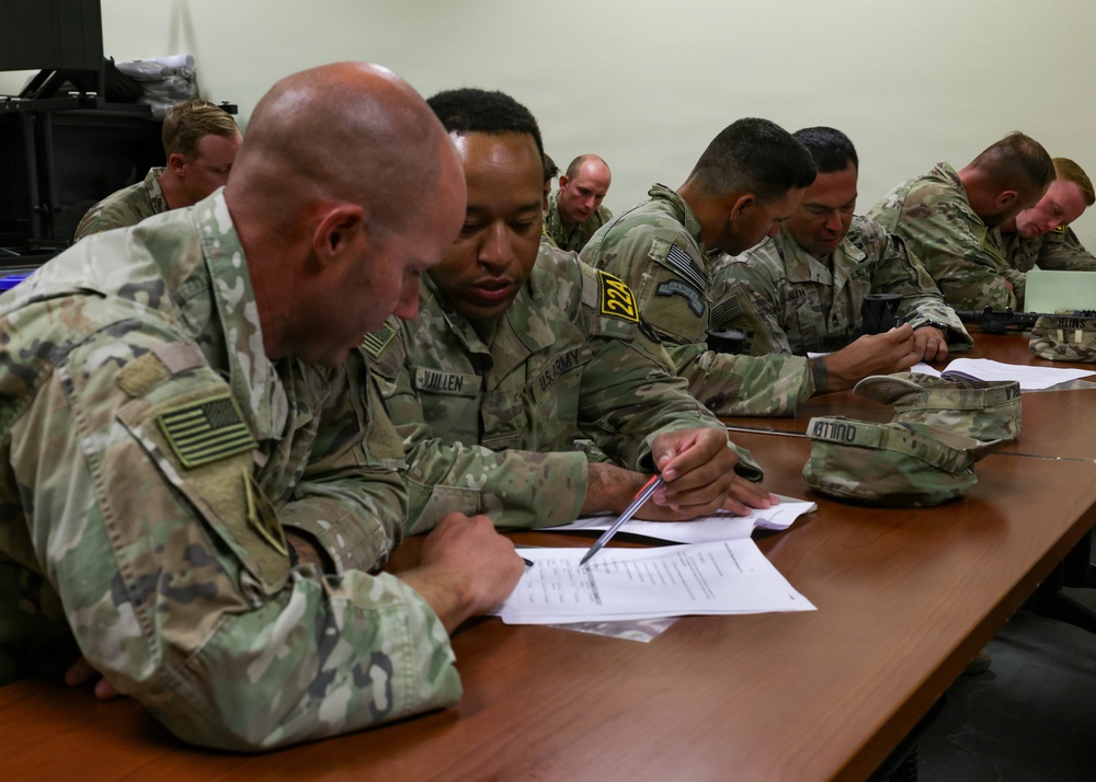 A group of U.S. Army soldiers in OCP (Operational Camouflage Pattern) uniforms are seated at a long wooden table engaged in a classroom or briefing session. The soldiers appear to be reviewing and filling out paperwork or documents. The setting appears to be an indoor military training or administrative facility.