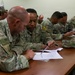 A group of U.S. Army soldiers in OCP (Operational Camouflage Pattern) uniforms are seated at a long wooden table engaged in a classroom or briefing session. The soldiers appear to be reviewing and filling out paperwork or documents. The setting appears to be an indoor military training or administrative facility.