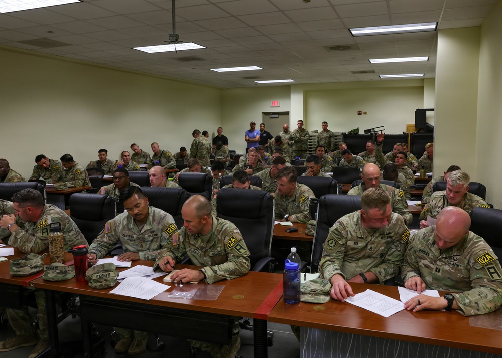 A large group of U.S. Army soldiers in OCP (Operational Camouflage Pattern) uniforms are seated in a tiered classroom or briefing room, reviewing documents and papers. The setting appears to be a formal military training or briefing session at a military installation. Soldiers are arranged in rows at desks, with name placards visible at the front tables.