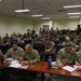 A large group of U.S. Army soldiers in OCP (Operational Camouflage Pattern) uniforms are seated in a tiered classroom or briefing room, reviewing documents and papers. The setting appears to be a formal military training or briefing session at a military installation. Soldiers are arranged in rows at desks, with name placards visible at the front tables.