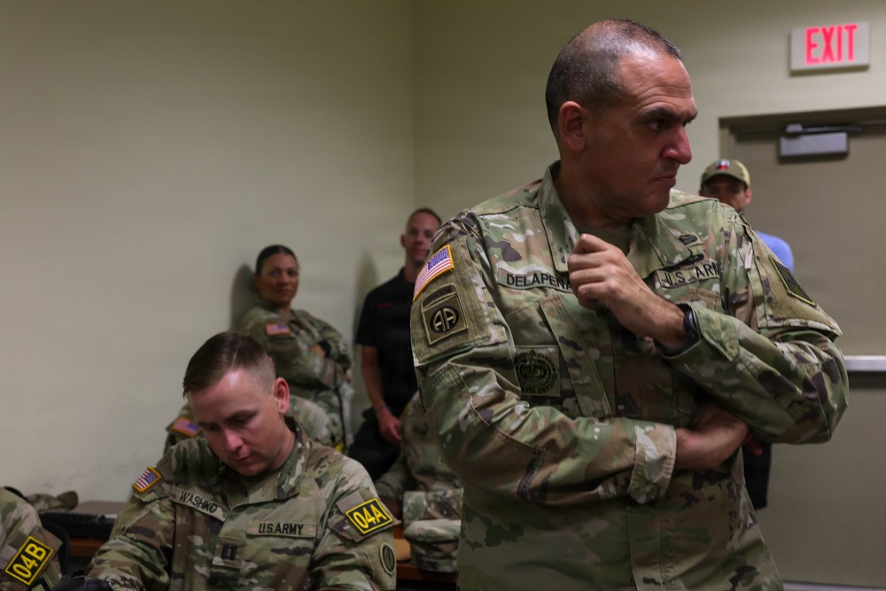 The image shows U.S. Army personnel in an indoor military setting, likely a briefing room or operations center. Two soldiers in the foreground are prominently visible wearing OCP (Operational Camouflage Pattern) uniforms, with name tapes 'DELAPENA' and 'WASHKO' legible. Several civilians and additional military personnel are visible in the background near an exit sign.