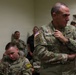 The image shows U.S. Army personnel in an indoor military setting, likely a briefing room or operations center. Two soldiers in the foreground are prominently visible wearing OCP (Operational Camouflage Pattern) uniforms, with name tapes 'DELAPENA' and 'WASHKO' legible. Several civilians and additional military personnel are visible in the background near an exit sign.