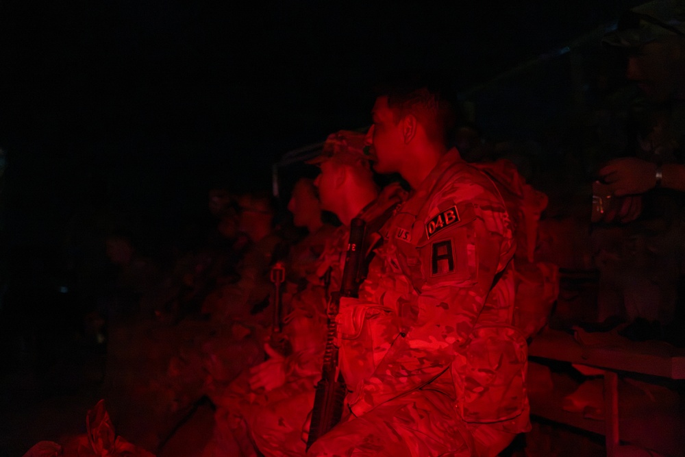 Two military personnel are illuminated by red light in an otherwise dark environment, wearing camouflage uniforms with visible patches and carrying weapons. The red lighting suggests a tactical or low-visibility operational setting, commonly used to preserve night vision. At least one soldier has visible unit patches including what appears to read 'U.S.' and 'DMB' with an 'A' patch.