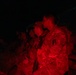 Two military personnel are illuminated by red light in an otherwise dark environment, wearing camouflage uniforms with visible patches and carrying weapons. The red lighting suggests a tactical or low-visibility operational setting, commonly used to preserve night vision. At least one soldier has visible unit patches including what appears to read 'U.S.' and 'DMB' with an 'A' patch.