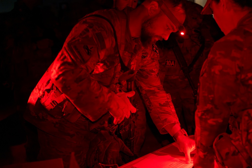 Military personnel in camouflage uniforms are gathered around a surface, illuminated by red light in a dark environment. At least one soldier has a visible 'ARMY' tape and a unit patch reading '10A' on their uniform. The red lighting suggests a tactical or night-operations setting where white light discipline is being maintained.
