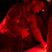 Military personnel in camouflage uniforms are gathered around a surface, illuminated by red light in a dark environment. At least one soldier has a visible 'ARMY' tape and a unit patch reading '10A' on their uniform. The red lighting suggests a tactical or night-operations setting where white light discipline is being maintained.