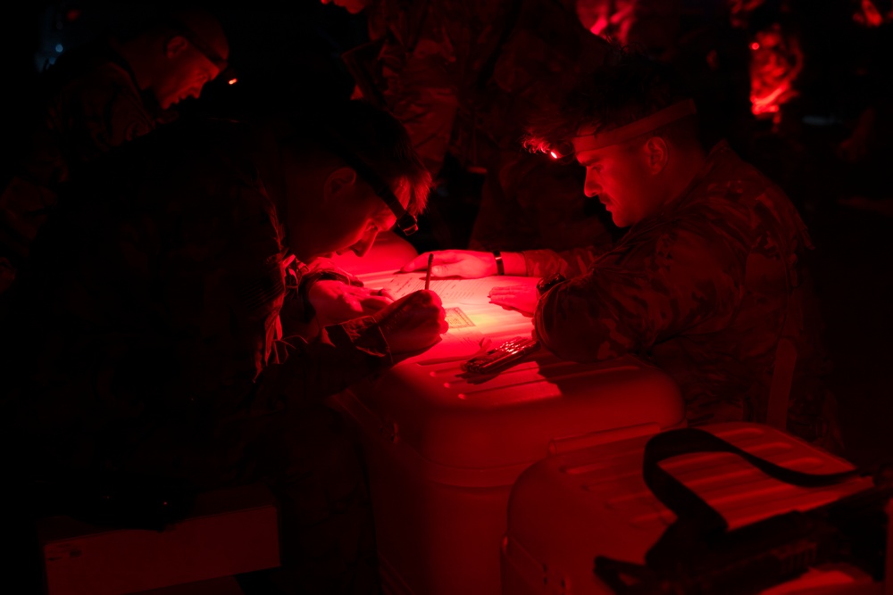 Military personnel are gathered around a table in near-total darkness, illuminated by red light, reviewing and writing on documents. The use of red light is consistent with tactical night operations, as red light preserves night vision. At least two individuals in camouflage uniforms are actively working on paperwork at what appears to be a field operations table.