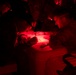 Military personnel are gathered around a table in near-total darkness, illuminated by red light, reviewing and writing on documents. The use of red light is consistent with tactical night operations, as red light preserves night vision. At least two individuals in camouflage uniforms are actively working on paperwork at what appears to be a field operations table.
