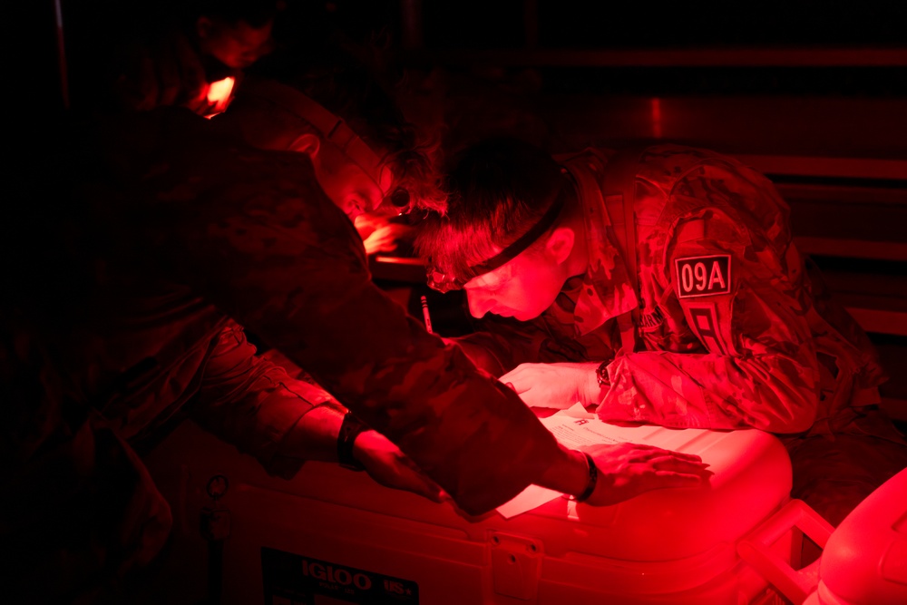 Military personnel are working in low-light conditions illuminated by red light, examining documents or paperwork placed on top of an Igloo cooler. At least two individuals in camouflage uniforms are visible, with one wearing a headlamp and a patch labeled '09A'. The red lighting suggests a tactical or field environment where white light discipline is being maintained.