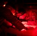 Military personnel are working in low-light conditions illuminated by red light, examining documents or paperwork placed on top of an Igloo cooler. At least two individuals in camouflage uniforms are visible, with one wearing a headlamp and a patch labeled '09A'. The red lighting suggests a tactical or field environment where white light discipline is being maintained.