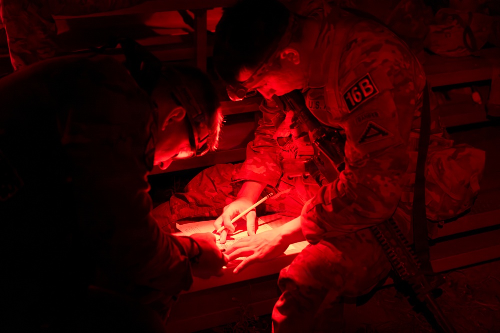Military personnel are gathered under red light conditions, reviewing what appears to be a map or document on a flat surface. The scene is illuminated entirely by red light, which is commonly used in tactical/night operations to preserve night vision. At least two soldiers in camouflage uniforms are visible, with one wearing a helmet patch marked '16B'.