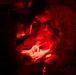 Military personnel are gathered under red light conditions, reviewing what appears to be a map or document on a flat surface. The scene is illuminated entirely by red light, which is commonly used in tactical/night operations to preserve night vision. At least two soldiers in camouflage uniforms are visible, with one wearing a helmet patch marked '16B'.