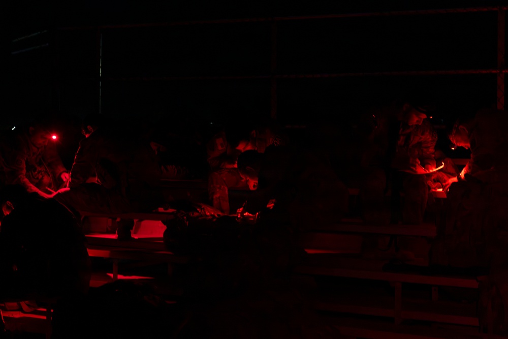 A very dark image illuminated primarily by red light sources, showing multiple individuals gathered around what appears to be a table or work surface. The red lighting suggests a tactical or night-operations environment, consistent with military or emergency personnel using red-light discipline to preserve night vision. Several people appear to be working on or examining materials on the surface in front of them.