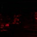 A very dark image illuminated primarily by red light sources, showing multiple individuals gathered around what appears to be a table or work surface. The red lighting suggests a tactical or night-operations environment, consistent with military or emergency personnel using red-light discipline to preserve night vision. Several people appear to be working on or examining materials on the surface in front of them.