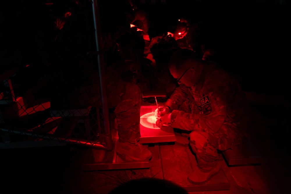 A military personnel in camouflage uniform is working under red light illumination at night, appearing to write or plot on a map or document. The scene is a nighttime military operations setting, likely a tactical operations center or field command post. Red light is used deliberately to preserve night vision while allowing work to continue in darkness.