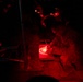 A military personnel in camouflage uniform is working under red light illumination at night, appearing to write or plot on a map or document. The scene is a nighttime military operations setting, likely a tactical operations center or field command post. Red light is used deliberately to preserve night vision while allowing work to continue in darkness.