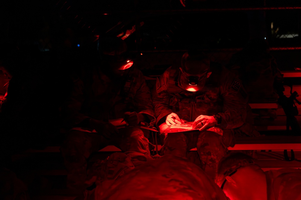 Military personnel are seated in what appears to be the interior of a transport aircraft or vehicle, illuminated by red lighting. One individual in the foreground is reviewing a document or map using a red-light source, a common practice to preserve night vision. The scene suggests a pre-mission briefing or in-transit review under tactical low-light conditions.
