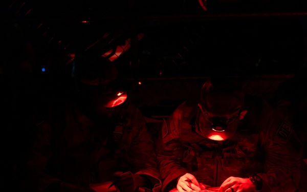 Military personnel are seated in what appears to be the interior of a transport aircraft or vehicle, illuminated by red lighting. One individual in the foreground is reviewing a document or map using a red-light source, a common practice to preserve night vision. The scene suggests a pre-mission briefing or in-transit review under tactical low-light conditions.
