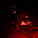 Military personnel are seated in what appears to be the interior of a transport aircraft or vehicle, illuminated by red lighting. One individual in the foreground is reviewing a document or map using a red-light source, a common practice to preserve night vision. The scene suggests a pre-mission briefing or in-transit review under tactical low-light conditions.