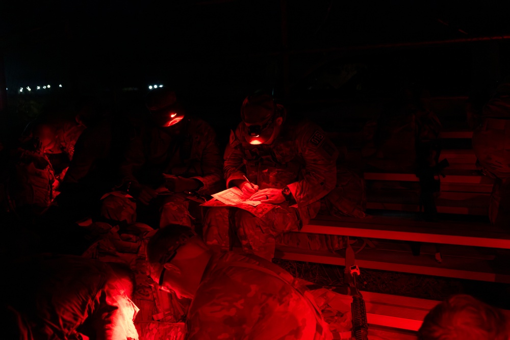 Military personnel are gathered at night, illuminated by red tactical lights, reviewing documents or maps. The soldiers appear to be in a briefing or planning session, seated on what appears to be a military vehicle or bench structure. Red light is used to preserve night vision while still allowing reading of materials.