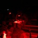 Military personnel are gathered at night, illuminated by red tactical lights, reviewing documents or maps. The soldiers appear to be in a briefing or planning session, seated on what appears to be a military vehicle or bench structure. Red light is used to preserve night vision while still allowing reading of materials.