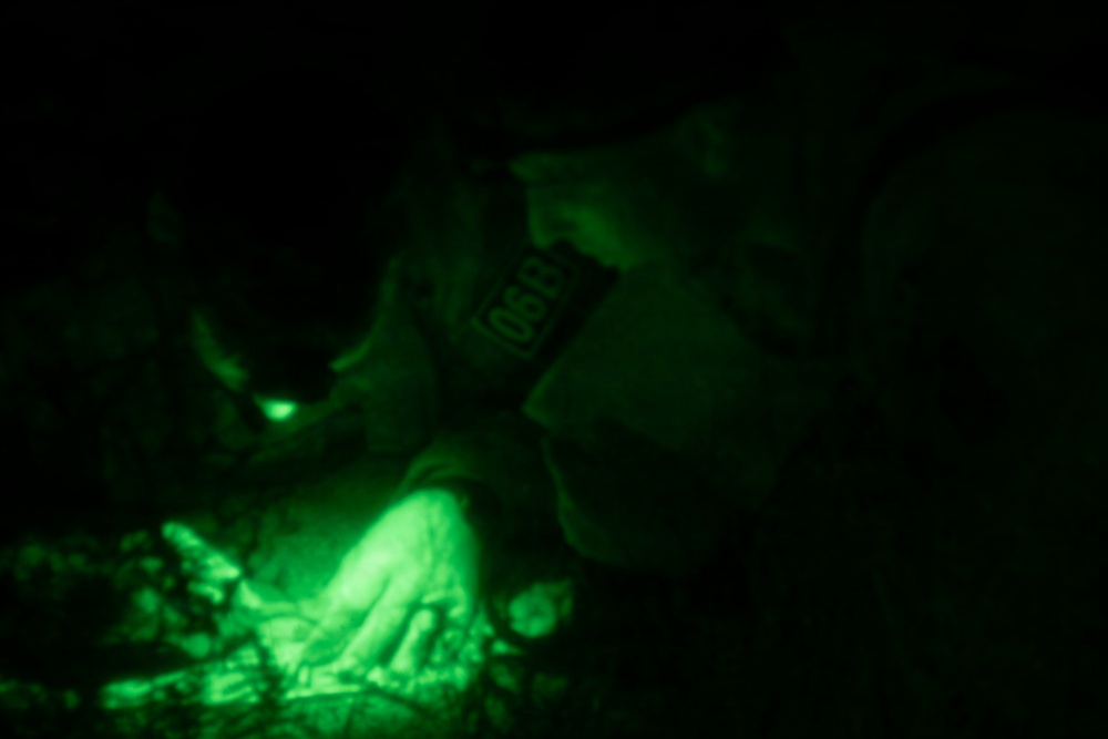 A person wearing military or tactical gear is working at ground level in complete darkness, illuminated only by a green light source. A patch is visible on their shoulder, appearing to read '068' or similar characters. The scene is captured under night vision or green-tinted lighting conditions.