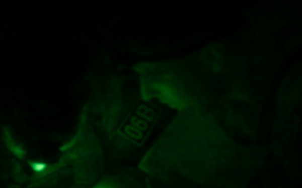 A person wearing military or tactical gear is working at ground level in complete darkness, illuminated only by a green light source. A patch is visible on their shoulder, appearing to read '068' or similar characters. The scene is captured under night vision or green-tinted lighting conditions.