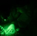 A person wearing military or tactical gear is working at ground level in complete darkness, illuminated only by a green light source. A patch is visible on their shoulder, appearing to read '068' or similar characters. The scene is captured under night vision or green-tinted lighting conditions.