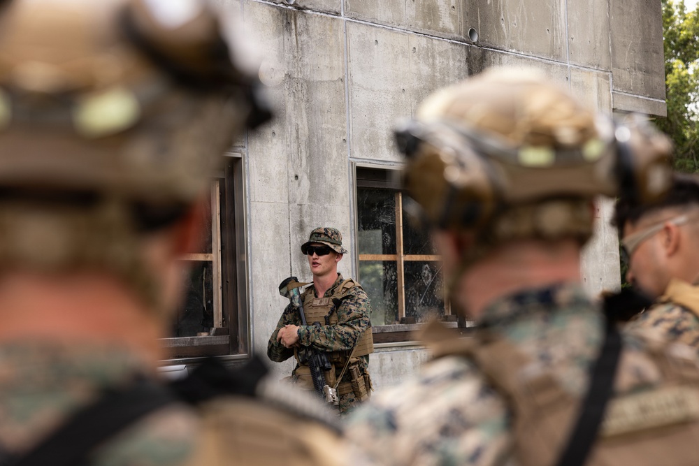A military instructor or non-commissioned officer in camouflage uniform and boonie hat addresses a group of soldiers or Marines in full combat gear. The setting appears to be a military training facility featuring a concrete structure with windows showing signs of simulated damage or weathering. The scene is consistent with a tactical training exercise or pre-mission briefing.