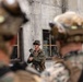 A military instructor or non-commissioned officer in camouflage uniform and boonie hat addresses a group of soldiers or Marines in full combat gear. The setting appears to be a military training facility featuring a concrete structure with windows showing signs of simulated damage or weathering. The scene is consistent with a tactical training exercise or pre-mission briefing.