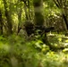 A soldier in full combat gear is positioned behind a tree in a dense forest, aiming a rifle in a tactical shooting stance. The individual is wearing digital camouflage uniform and a helmet with attached equipment. The setting appears to be a woodland training exercise or operational patrol in a lush, green forest environment.