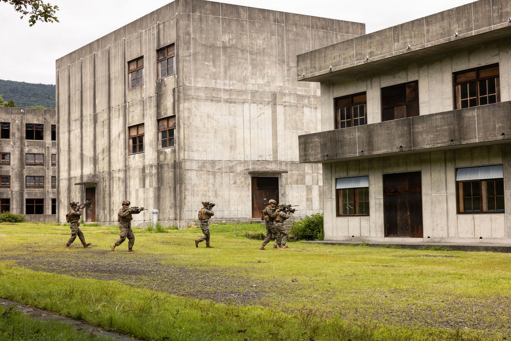 Three armed military personnel in camouflage uniforms are conducting what appears to be a tactical training exercise in an urban warfare simulation environment. They are moving in formation across an open grassy area toward concrete multi-story buildings that appear to be purpose-built training structures. The soldiers are carrying rifles in a ready position, suggesting a building-clearing or assault drill.