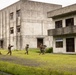 Three armed military personnel in camouflage uniforms are conducting what appears to be a tactical training exercise in an urban warfare simulation environment. They are moving in formation across an open grassy area toward concrete multi-story buildings that appear to be purpose-built training structures. The soldiers are carrying rifles in a ready position, suggesting a building-clearing or assault drill.