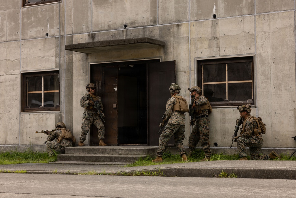 A group of military personnel in full combat gear are conducting a building entry/clearing exercise at what appears to be a training facility. Five soldiers in woodland/digital camouflage uniforms are positioned around the entrance of a concrete structure in a tactical stack formation. The scene depicts a Military Operations in Urban Terrain (MOUT) training drill.