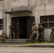 A group of military personnel in full combat gear are conducting a building entry/clearing exercise at what appears to be a training facility. Five soldiers in woodland/digital camouflage uniforms are positioned around the entrance of a concrete structure in a tactical stack formation. The scene depicts a Military Operations in Urban Terrain (MOUT) training drill.