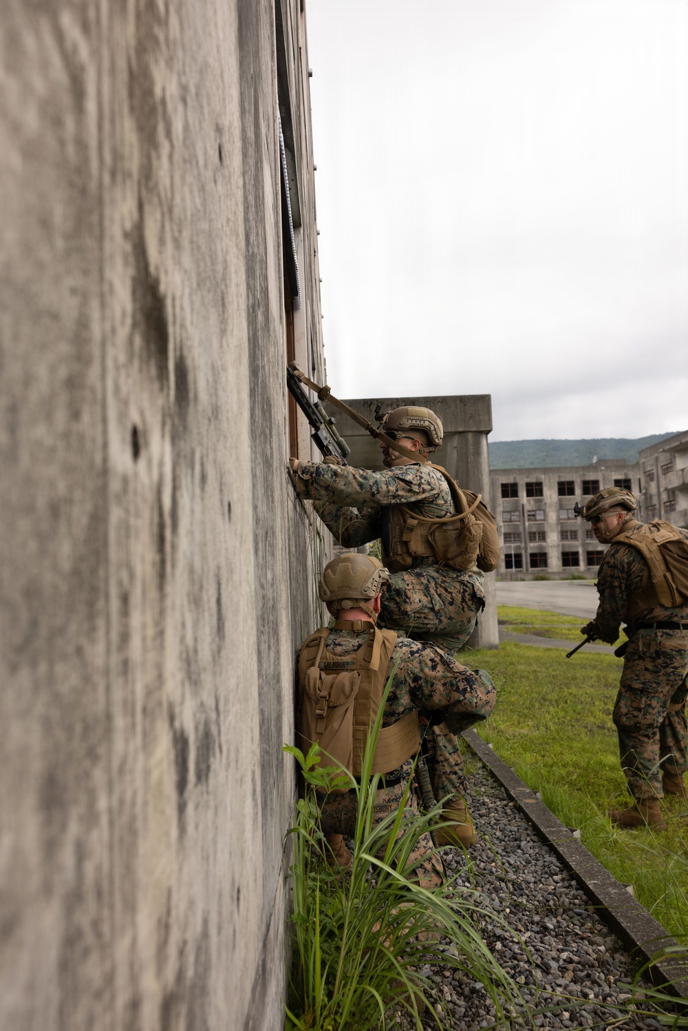 Three military personnel in U.S. Marine Corps digital camouflage uniforms are conducting urban operations training at what appears to be an abandoned or mock facility. Two Marines are stacked against a concrete wall, with one peering through or over a window opening while holding a rifle, and a third Marine stands nearby providing security. The setting includes deteriorating concrete buildings and overgrown vegetation, consistent with a military training environment.