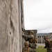 Three military personnel in U.S. Marine Corps digital camouflage uniforms are conducting urban operations training at what appears to be an abandoned or mock facility. Two Marines are stacked against a concrete wall, with one peering through or over a window opening while holding a rifle, and a third Marine stands nearby providing security. The setting includes deteriorating concrete buildings and overgrown vegetation, consistent with a military training environment.