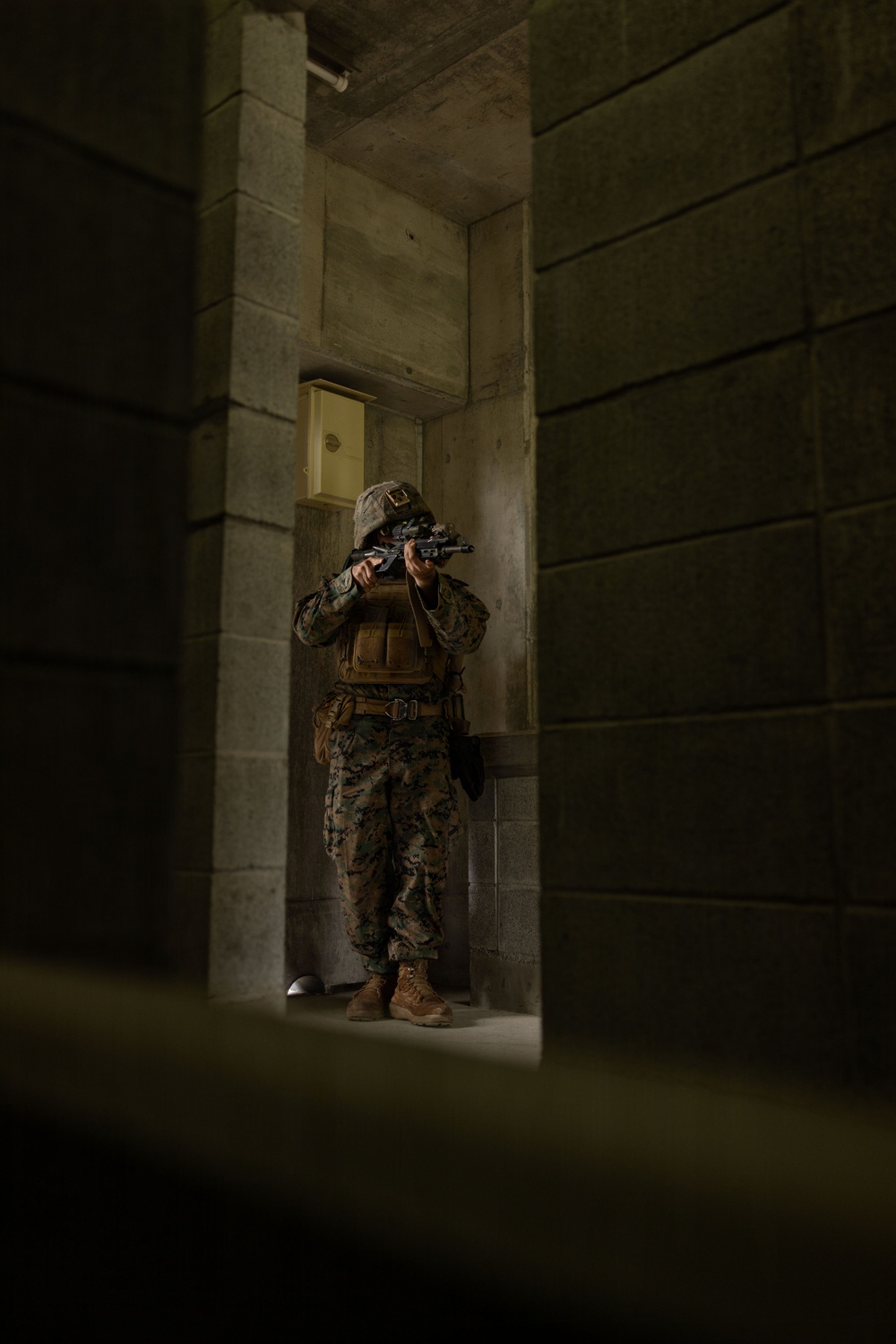 A military service member in full combat gear is photographed through a narrow corridor or doorway in a concrete structure. The soldier is in an aiming stance with a rifle raised to eye level, suggesting a training exercise or tactical operation in an urban/building environment. The image is captured from a distance through an architectural gap, creating a framed, dramatic composition.