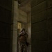 A military service member in full combat gear is photographed through a narrow corridor or doorway in a concrete structure. The soldier is in an aiming stance with a rifle raised to eye level, suggesting a training exercise or tactical operation in an urban/building environment. The image is captured from a distance through an architectural gap, creating a framed, dramatic composition.