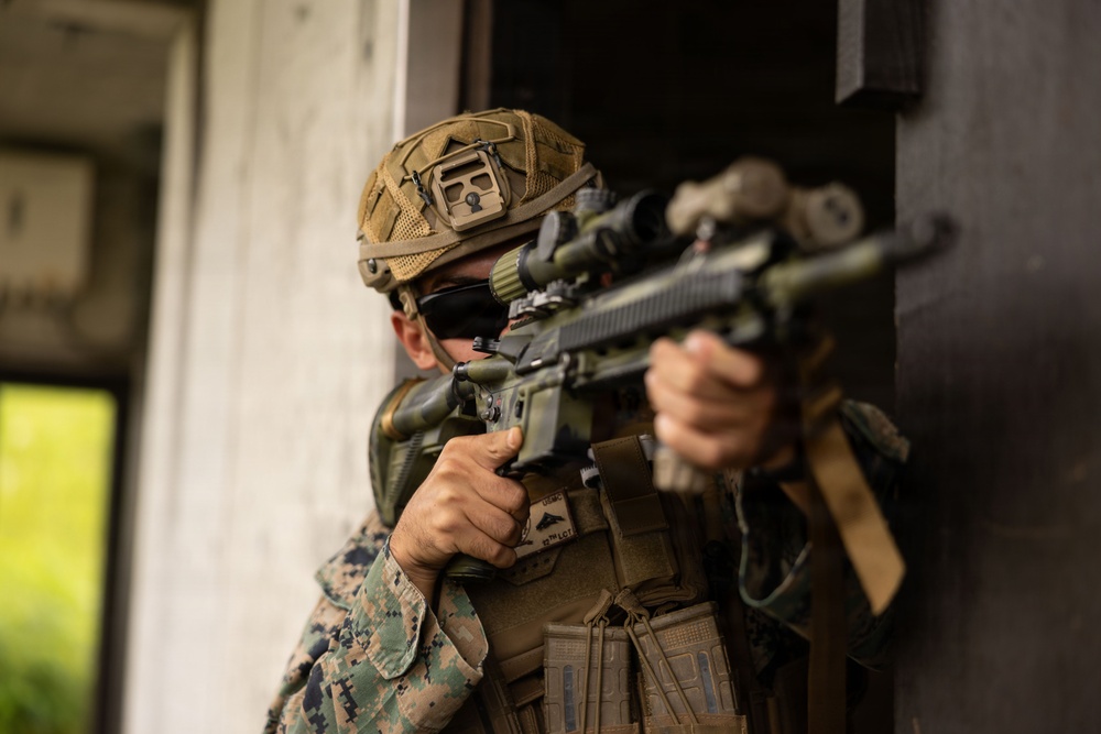 A military service member in MARPAT (Marine pattern) digital camouflage uniform is positioned at a doorway or building corner, aiming a scoped precision rifle. The individual is wearing full tactical gear including a helmet with rail-mounted accessories, a face covering, and a plate carrier vest. The setting appears to be an urban training environment or military exercise.