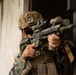 A military service member in MARPAT (Marine pattern) digital camouflage uniform is positioned at a doorway or building corner, aiming a scoped precision rifle. The individual is wearing full tactical gear including a helmet with rail-mounted accessories, a face covering, and a plate carrier vest. The setting appears to be an urban training environment or military exercise.