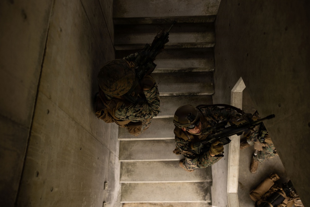 An overhead (top-down) photograph shows two or three armed military personnel ascending a concrete stairwell. The personnel are wearing camouflage uniforms, tactical gear, and helmets, and are carrying assault rifles in a tactical clearing posture. The image appears to depict a military training exercise in an urban operations or close-quarters battle (CQB) environment.