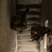 An overhead (top-down) photograph shows two or three armed military personnel ascending a concrete stairwell. The personnel are wearing camouflage uniforms, tactical gear, and helmets, and are carrying assault rifles in a tactical clearing posture. The image appears to depict a military training exercise in an urban operations or close-quarters battle (CQB) environment.