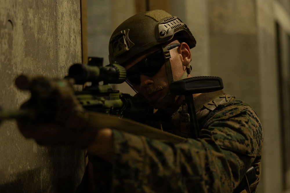 A military service member in full tactical gear is positioned against a concrete wall, aiming a rifle with an optical scope. The individual is wearing MARPAT (Marine pattern) camouflage uniform, a tactical helmet with rail-mounted accessories, and ballistic eyewear. The image appears to depict a tactical training or operational scenario in an urban or structured environment.