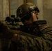 A military service member in full tactical gear is positioned against a concrete wall, aiming a rifle with an optical scope. The individual is wearing MARPAT (Marine pattern) camouflage uniform, a tactical helmet with rail-mounted accessories, and ballistic eyewear. The image appears to depict a tactical training or operational scenario in an urban or structured environment.