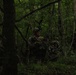 A group of armed military personnel in camouflage uniforms are conducting a patrol or tactical movement through a dense forest environment. The soldiers are equipped with rifles, tactical helmets, and protective eyewear, suggesting a training exercise or operational mission. The low-light, heavily wooded setting creates a dark and concealed atmosphere.