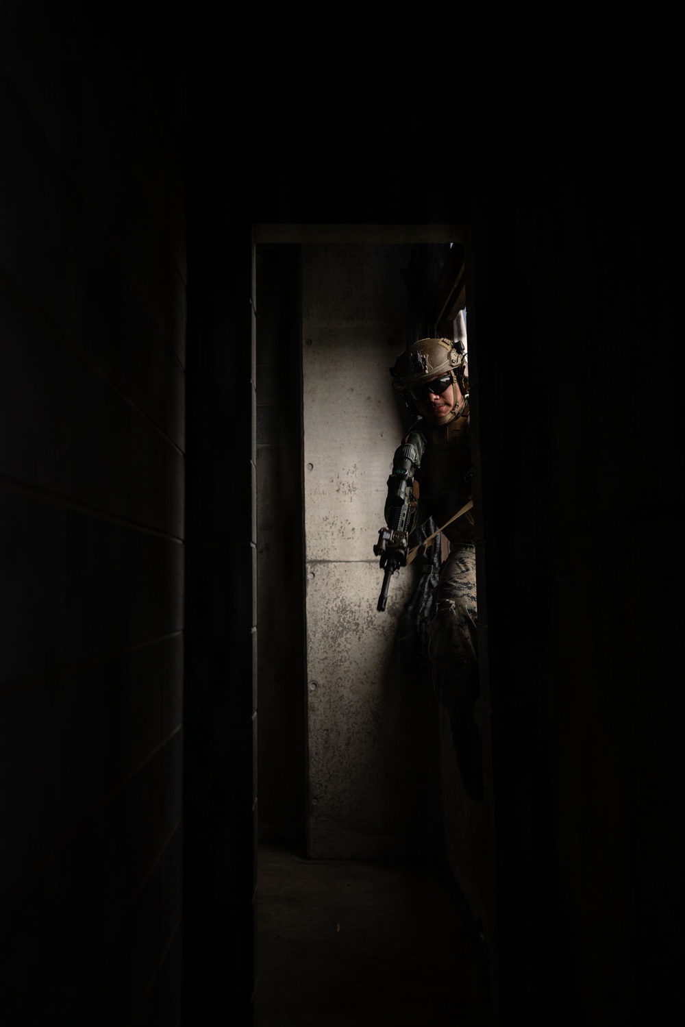 A heavily equipped military or law enforcement operator is visible through a doorway in a darkened structure, pressing against a concrete wall in a tactical stance. The individual is wearing full combat gear including a helmet, body armor, tactical gloves, and is holding a rifle. The dramatic lighting creates a high-contrast scene consistent with military training or operational photography.