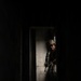 A heavily equipped military or law enforcement operator is visible through a doorway in a darkened structure, pressing against a concrete wall in a tactical stance. The individual is wearing full combat gear including a helmet, body armor, tactical gloves, and is holding a rifle. The dramatic lighting creates a high-contrast scene consistent with military training or operational photography.